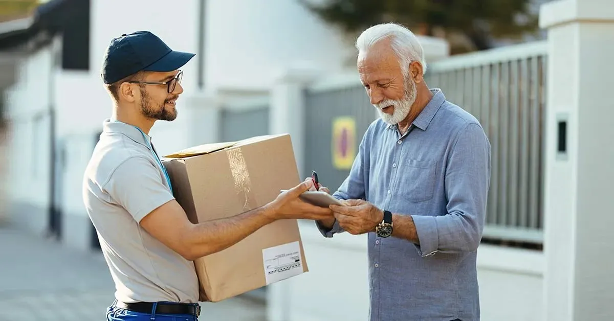 A courier in a cap handing a cardboard parcel to a customer at his front gate