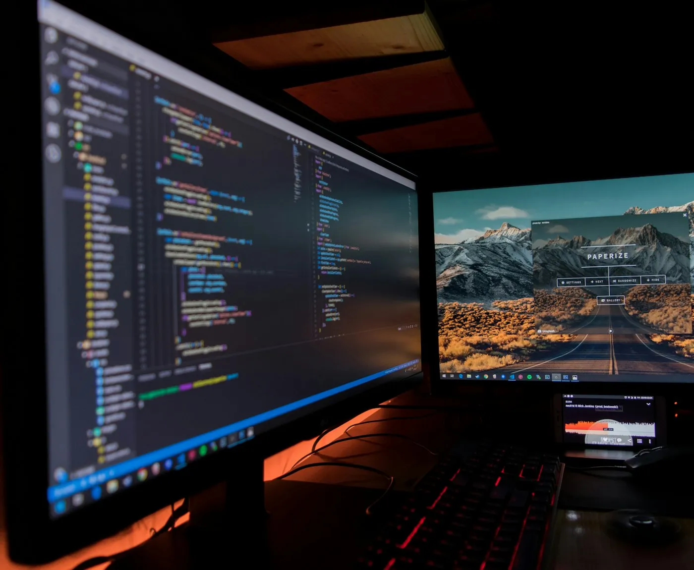 Two monitors with code and a mountain road photo on a dark desk