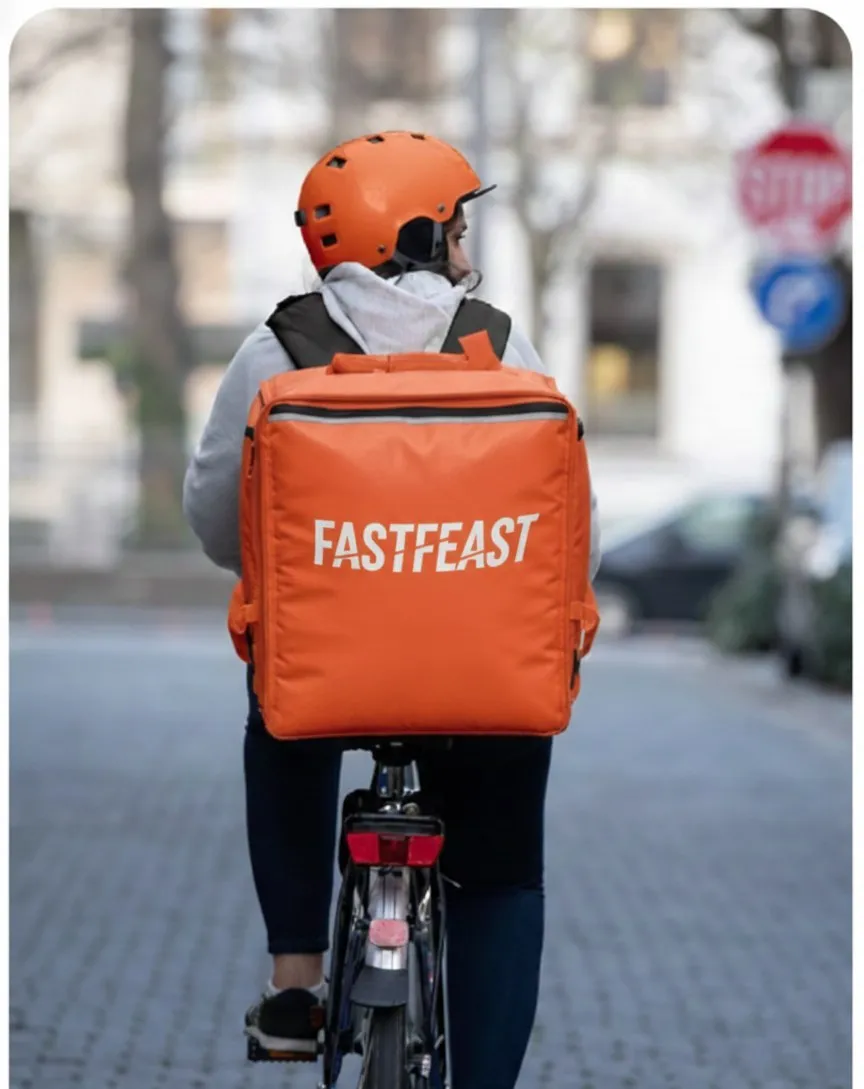 A delivery rider on a bicycle carrying an orange FastFeast delivery backpack down a city street